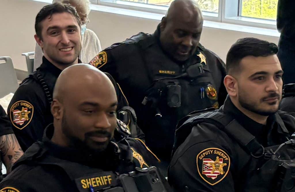 Vajusi, a man with short, dark-brown hair, wears a sheriff's department uniform. He sits next to three other men, also wearing sheriff's department uniforms, in a room with windows with sunlight coming through.