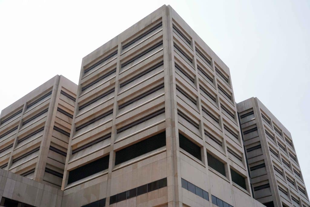 A photo, taken from a low angle, shows the outside of a light brown tower with narrow, horizontal windows. The sky is visible above the building.