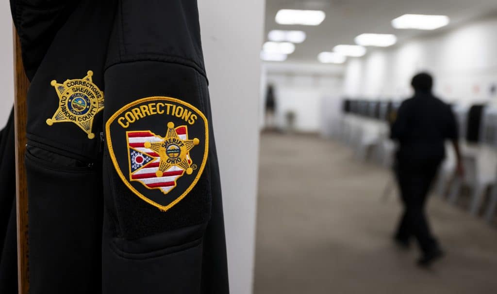 On the left, in focus, hangs a jacket with a patch that says “Corrections.” The design includes the state flag in the shape of Ohio with a sheriff’s star on top. Another star-shaped patch says “Corrections, Sheriff, Cuyahoga County.” On the right, out of focus, a person walks through the video visitation room of the Cuyahoga County Justice Complex.
