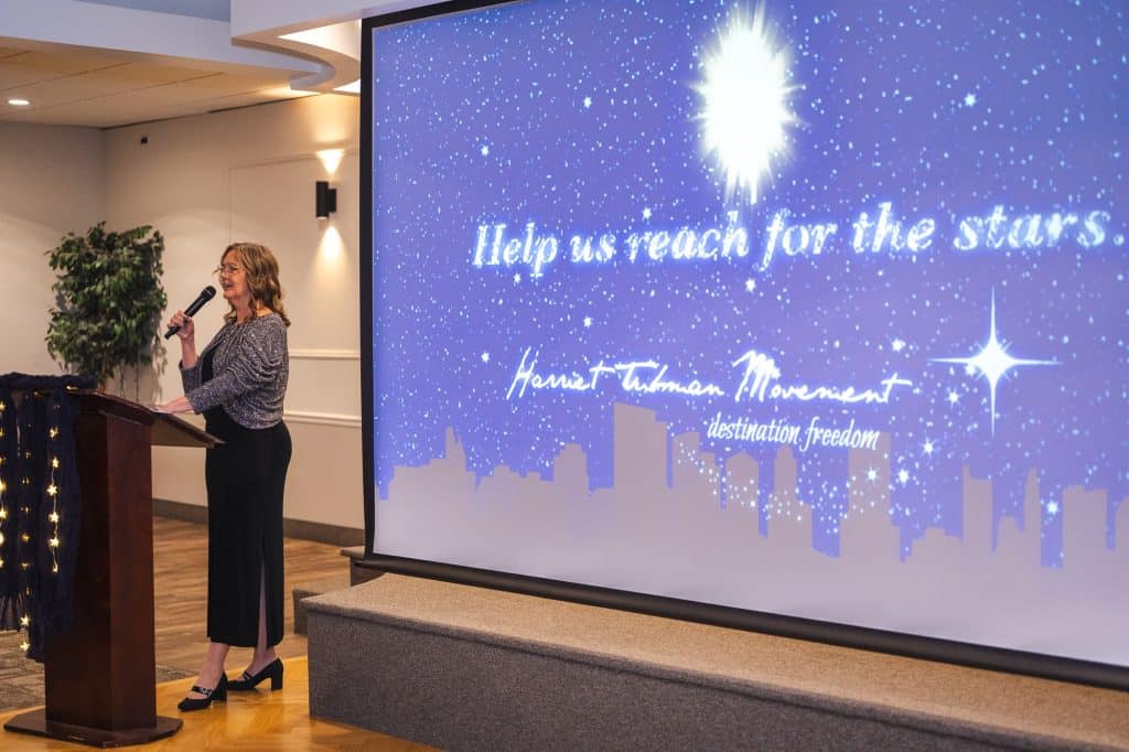 Laura Bartchak, a white woman, stands in front of a screen and delivers a speech. 