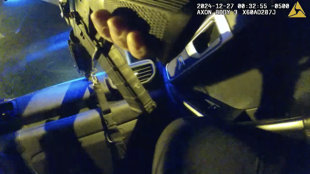 A screenshot of bodycam footage shows a person’s hand holding an assault rifle inside a vehicle at night.