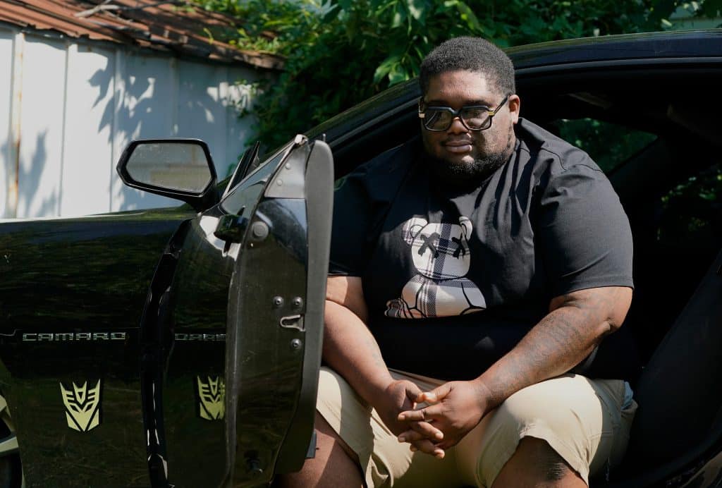 Henry Ray, a Black man, wears a black T-shirt, khaki shorts and sunglasses while sitting in his Camaro with the door open.