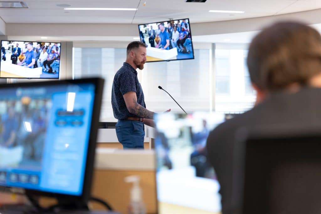 James DeCredico, a White man wearing a blue short-sleeved shirt and blue pants, stands at a podium in a county council meeting. There are two monitors showing the proceedings behind him.