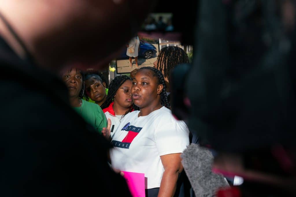 Cearria Elder, a Black woman wearing a white Tommy Hilfiger T-shirt, is visible through the space between a person and a video camera.
