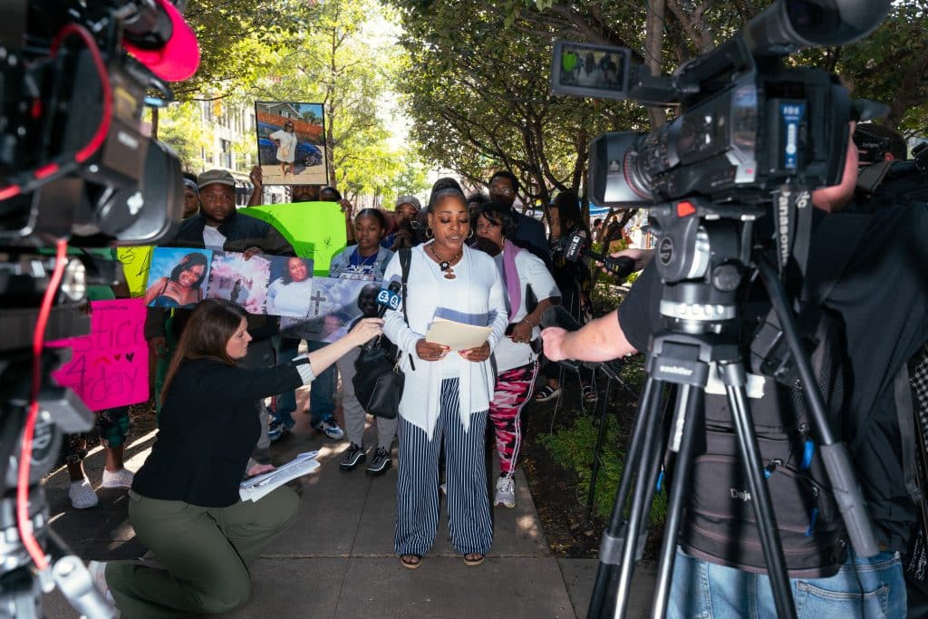 LaTonya Goldsby, a Black woman wearing a white cardigan, white shirt and striped pants, speaks at an outdoor press conference. A group of people stands behind her, holding family photos. News video cameras are in the foreground.