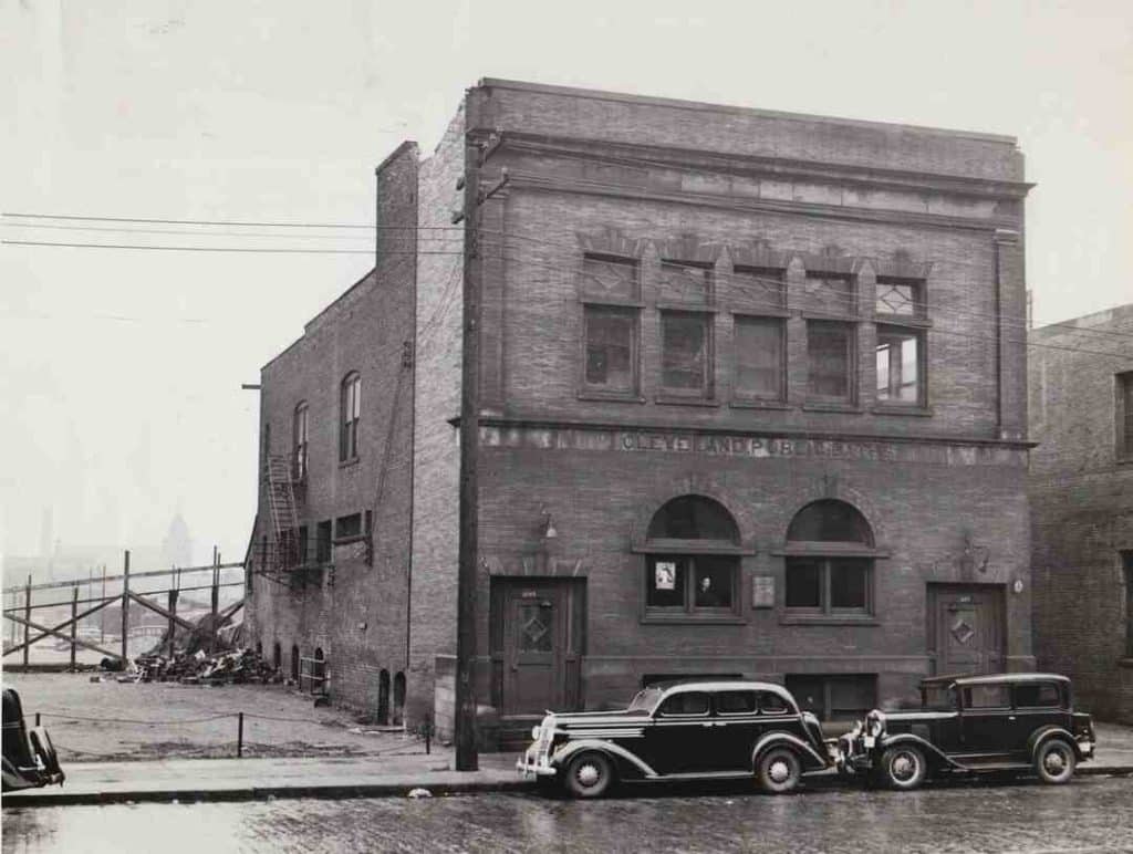 A black-and-white photo of a brick building with two old automobiles parked in front of the facade. 