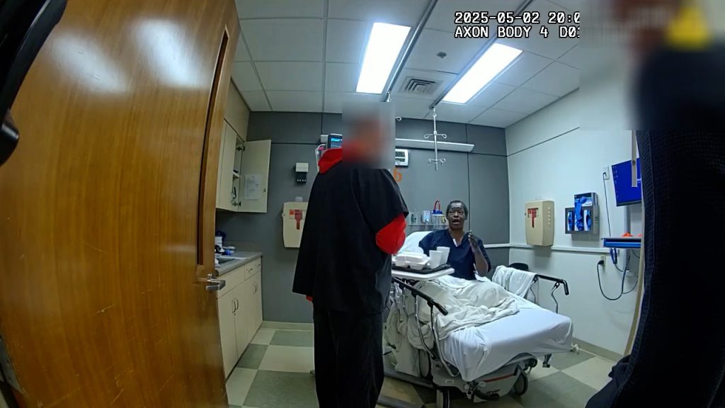 Tasha Grant, a Black woman with close-cropped hair and wearing a dark blue shirt, sits on a hospital bed and can be seen talking with a medical staff member who stands a few feet away to the left of the frame, wearing dark blue scrubs over a red hooded sweatshirt. His face is blurred out.