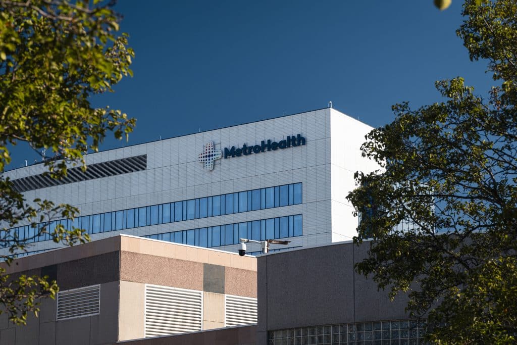 A photo of the MetroHealth Medical Center. The building is white and rectangular, has two lines of visible windows, and the word “MetroHealth” is on the top right of the building. In front of that building is a lower beige/orange-toned building.