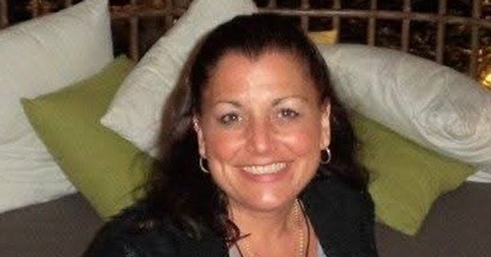 A smiling woman with light-toned skin and medium-length dark brown hair sits in front of some white and pale green pillows.