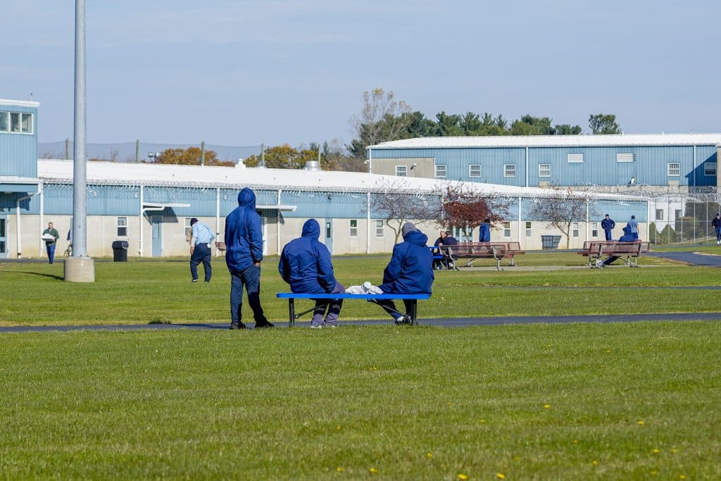 A photo shows three incarcerated people dressed in blue uniforms in the foreground, outdoors, but on prison grounds. Their faces are not visible, and two of them are sitting on a bench, and one is standing to the left. Two buildings are visible behind the three men, which are pale blue and white, and there are a few other people visible in the background. In the distance, netting along the fence line is visible.