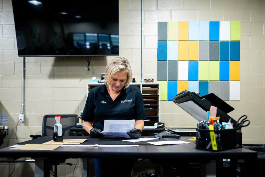 A photo shows Karen Johnson, a White woman with blonde hair, wearing a black T-shirt and gloves, holding a piece of mail in a mail processing center.