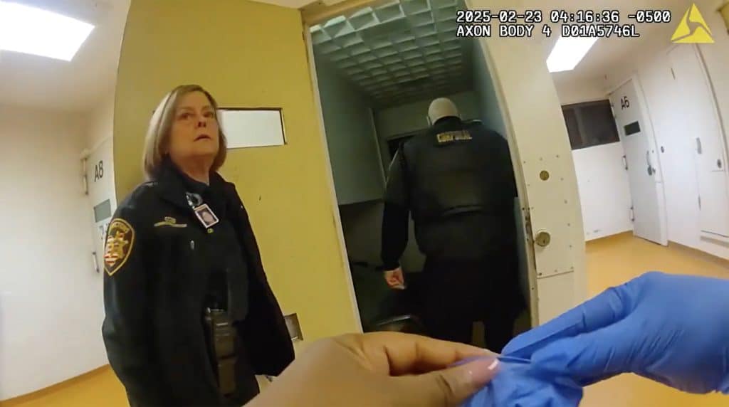 A still from a bodycam shows corrections officers outside Jennifer Wade's cell. Her body is not visible. In the foreground is a person putting on rubber gloves.