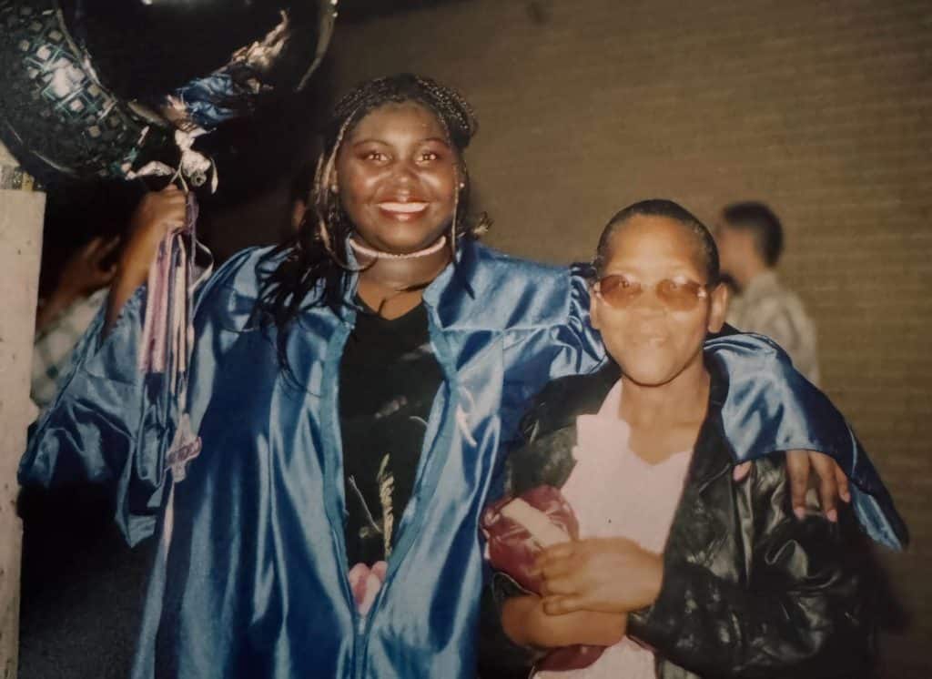 A photo shows Jennifer Wade, a Black woman wearing braids and a blue graduation gown, holding balloons and with her arm around her grandmother, Florine Wade, a Black woman wearing a black coat and a white shirt.  