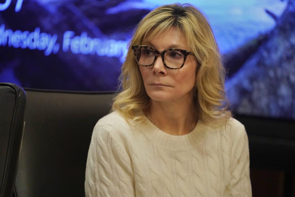 A photo shows Leslie Ann Celebrezze, a White woman with blond hair, wearing an ivory sweater, sitting in front of a large monitor with blue mountains and a lake on it.